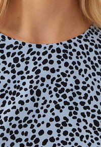 Tissu bleu clair présentant un motif texturé de petits pois noirs, avec un col rond et une finition lisse.