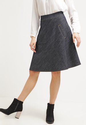 Vrouw draagt een donker denim A-lijn rok met zijzakken, gecombineerd met een witte blouse met lange mouwen en zwarte enkellaarsjes met blokhakken.