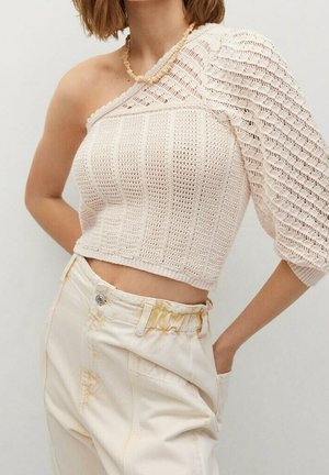 Mujer con un top beige de un solo hombro de crochet con manga abullonada y pantalones crema de cintura alta con cinturilla fruncida, con la mano en el bolsillo.