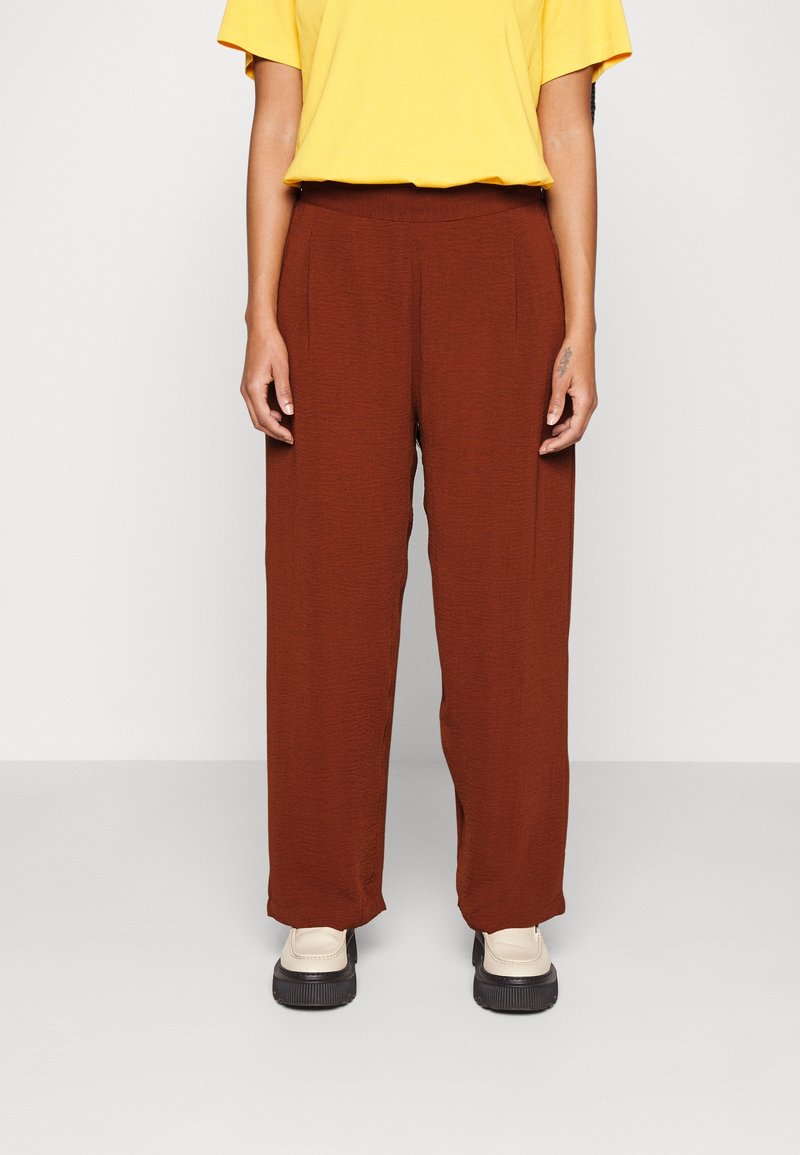Personne portant un pantalon marron ample, un haut jaune à manches courtes et des chaussures compensées crème, debout devant un fond uni.