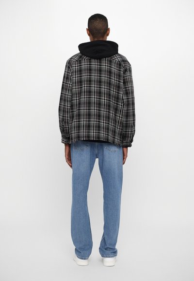 Veste en flanelle à carreaux noir et gris avec capuche, portée sur un haut noir, associée à un jean droit bleu clair et des baskets blanches.
