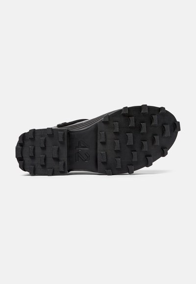 Suola di una scarpa sportiva nera con tacchetti profondi e multidirezionali per la trazione, superficie texturizzata e branding in rilievo al centro.