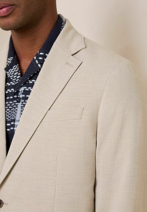 Mann trägt eine strukturierte beige Anzugjacke über einem schwarzen gemusterten Hemd, dargestellt vom Hals bis zur Mitte des Oberkörpers vor einfachem Hintergrund.