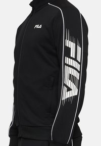 Chaqueta atlética negra con un collar alto, franjas laterales blancas y un gran logo de FILA en la manga. Hecha de un tejido suave y texturizado.