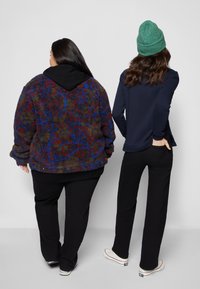 Duas figuras estão de costas uma para a outra: uma veste uma jaqueta felpuda e multicolorida com capuz preto, e a outra usa uma blusa azul-marinho e um gorro verde.