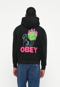 Hanorac negru cu un grafic ce prezintă o figură care ține un glob cu flăcări, textul îndrăzneț "OBEY" în roz și contururi albastre pe design.