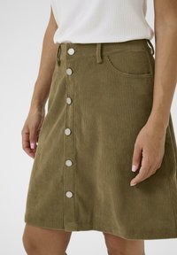 Olijfgroene corduroy rok met zilveren knopen aan de voorkant, gedragen met een witte ribgebreide mouwloze top, handen zichtbaar aan de zijkanten.