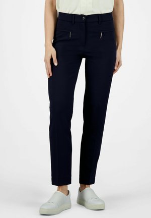 Pantalons navy à la texture lisse, présentant une coupe slim, des poches zippées à l'avant et une fermeture à bouton. Associés à des chaussures de couleur claire.