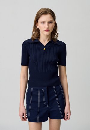 Top a coste navy a maniche corte con colletto, dotato di un bottone circolare dorato. Abbinato a pantaloni corti navy con dettagli di cucitura a contrasto.