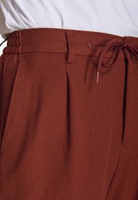Brune teksturerede joggerbukser med snor i taljen, med folder og en glat overflade. Egnet til hverdagsbrug.