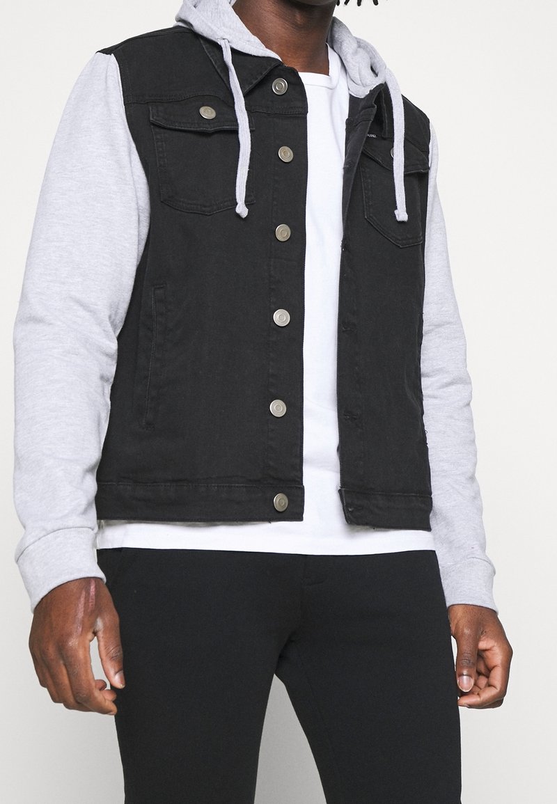 Czarna kamizelka jeansowa z szarą bluzą z kapturem, wyposażona w dwie kieszenie na piersi i zapięcia na zatrzaski. Noszona na białej koszulce i czarnych spodniach.