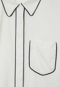 Weißes Shirt vorne mit spitzem Kragen, vertikalem Knopfverschluss und Brusttasche, alles mit dünnem schwarzem Piping eingefasst.