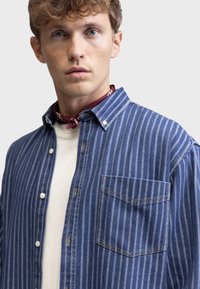 Blue striped denim shirt with a button-down collar, left chest pocket, and contrasting stitching. Worn over a light-colored t-shirt.