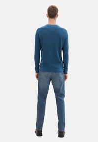Jersey de punto azul con puños acanalados, combinado con vaqueros de denim azul claro y zapatos negros, visto desde la parte de atrás contra un fondo liso.