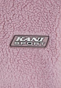Tejido de forro polar de color rosa claro con superficie texturizada y logotipo de KANI SPORT en negro y blanco en una etiqueta rectangular.