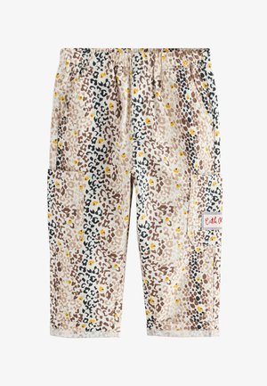 Jong kind staat met het gezicht naar een bloemenmuur, draagt een wit T-shirt, een broek met patroon en beige schoenen op een geometrisch betegelde vloer.