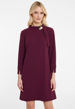 Robe droite bordeaux en tissu doux, avec des manches longues, des fentes sur les côtés et un col rond avec un accent doré près du col.