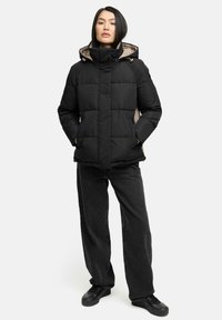 Zwarte pufferjas met een hoge kraag en een beige voering, voorzien van gestikte panelen, zijzakken en een capuchon met een trekkoord. Gecombineerd met zwarte jeans.