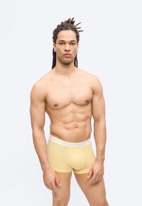 Άνδρας χωρίς πουκάμισο με dreadlocks φορώντας ανοιχτόκίτρινα μποξέρ στέκεται μπροστά σε απλό φωτεινό φόντο, κοιτάζοντας προς τα εμπρός με ουδέτερη έκφραση.