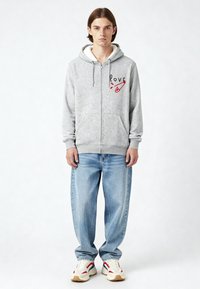 Felpa grigia con zip, tasche frontali, cappuccio foderato in pile e testo grafico rosso. Abbinata a jeans baggy blu chiaro e sneakers multicolori.