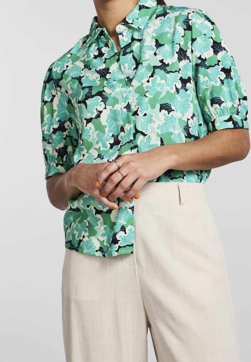 Chemise verte à manches courtes présentant un motif floral aux nuances variées de vert et de noir, associée à un pantalon de couleur claire.