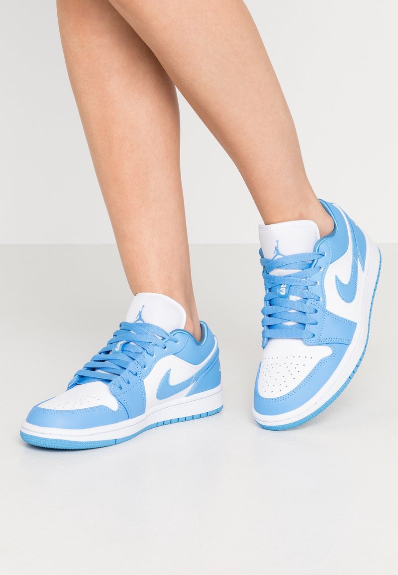 Ben bärande blå och vita Nike Air Jordan-sneakers med snören, stående på ett enfärgat ljust golv mot en enkel bakgrund.