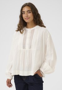 Femeie cu păr buclat, purtând o bluză albă largă cu detalii din dantelă și pliuri, pantaloni închiși la culoare și o brățară din argint, stând în fața unui fundal simplu.