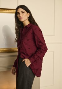 Bordeaux broderet bluse med krave og lange ærmer, prydet med blomsterdesigns og en løs pasform, parret med mørke stribede bukser.