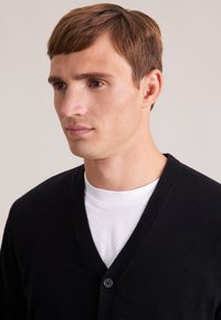 Cardigan noir à col en V en tricot, avec une fermeture à un bouton, porté sur un t-shirt blanc, montrant un gros plan de l'encolure et de l'ajustement.