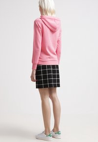 Sweat à capuche rose avec cordon de serrage et manches longues, associé à une jupe à carreaux noire et blanche. La tenue est complétée par des baskets blanches avec des accents verts.