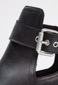 Czarny skórzany but z błyszczącą srebrną klamrą, z wyciętym wzorem i widocznym szwem wzdłuż krawędzi. Gładka tekstura.