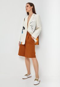 White blazer with abstract color prints, paired with brown shorts. The shoes are white with a gold chain accent. Smooth texture, tailored fit.