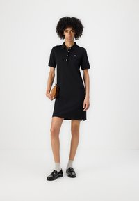 Rochie polo neagră într-un stil ajustat, cu mâneci scurte, placket cu trei butoane și logo brodat. Purtați cu șosete gri și mocasini negri.