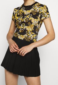 Czarna koszulka z żółtym, ozdobnym wzorem i napisem, w zestawie z plisowaną czarną spódnicą. Góra ma krótkie rękawy i okrągły dekolt.