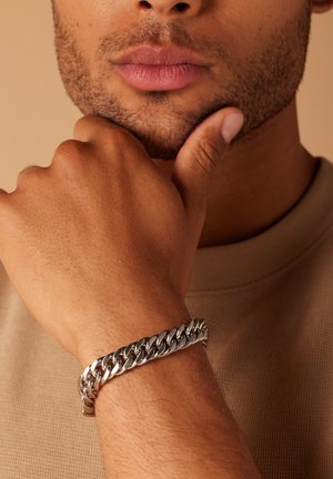 Silver chain bracelet on a wrist, featuring interlocking links with a polished finish. Background is soft beige fabric texture.