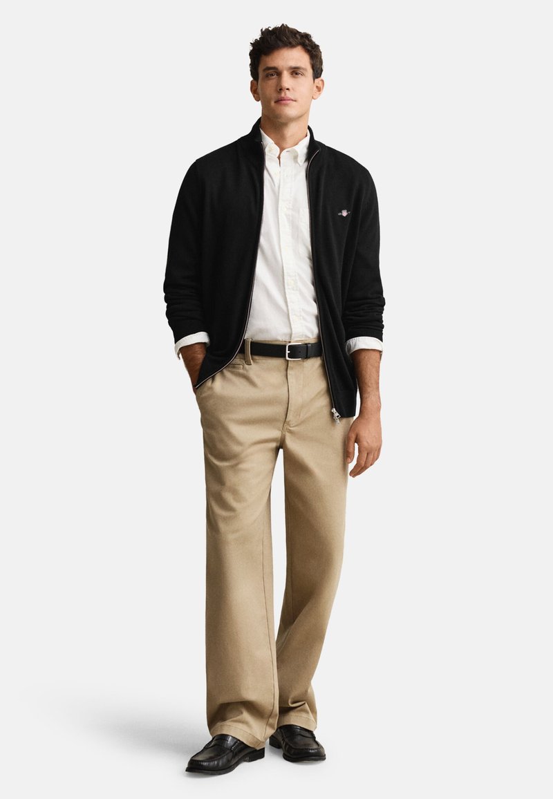 Casaco preto com fecho de correr por cima de uma camisa branca, combinado com calças bege, cinto preto e sapatos pretos. Tecido liso, corte ajustado e visual casual.