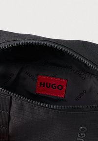 Μαύρη τσάντα με φερμουάρ, εσωτερικά εμφανίζεται μια κόκκινη ορθογώνια ετικέτα με την επιγραφή "HUGO" σε μαύρα γράμματα ραμμένη μέσα στην επένδυση.