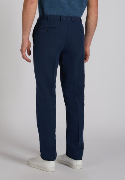 Mężczyzna w granatowych spodniach i białych trampkach stoi tyłem do kamery na jednolitym tle.