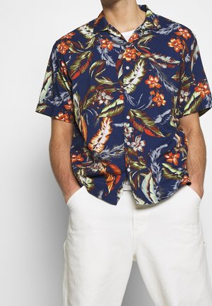 Camicia a maniche corte, con bottoni, di tessuto blu navy con vivaci motivi floreali e di foglie in rosso, arancione e verde. Indossata con pantaloni bianchi.