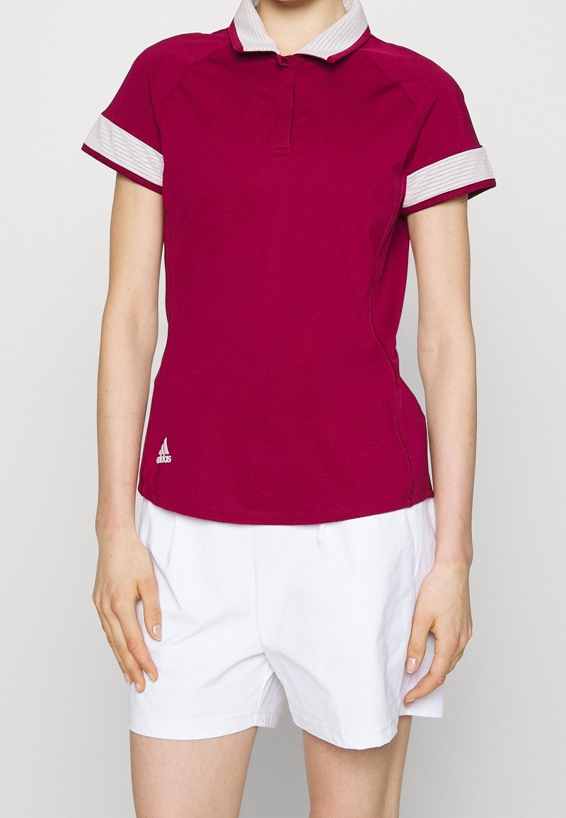 Personne portant un polo bordeaux à manches courtes avec col blanc et bordure blanche aux manches, associé à un short blanc.