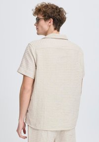 Camisa de manga corta color beige con un patrón texturizado, cuello abotonado y dobladillo recto, que presenta una etiqueta de marca sutil en el costado.