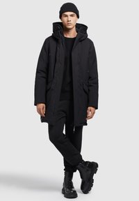 Sort lang parka-jakke med hætte, lavet af et glat materiale. Har lynlåslommer i siderne og justerbare manchetter. Bæres over en sort sweater og bukser.