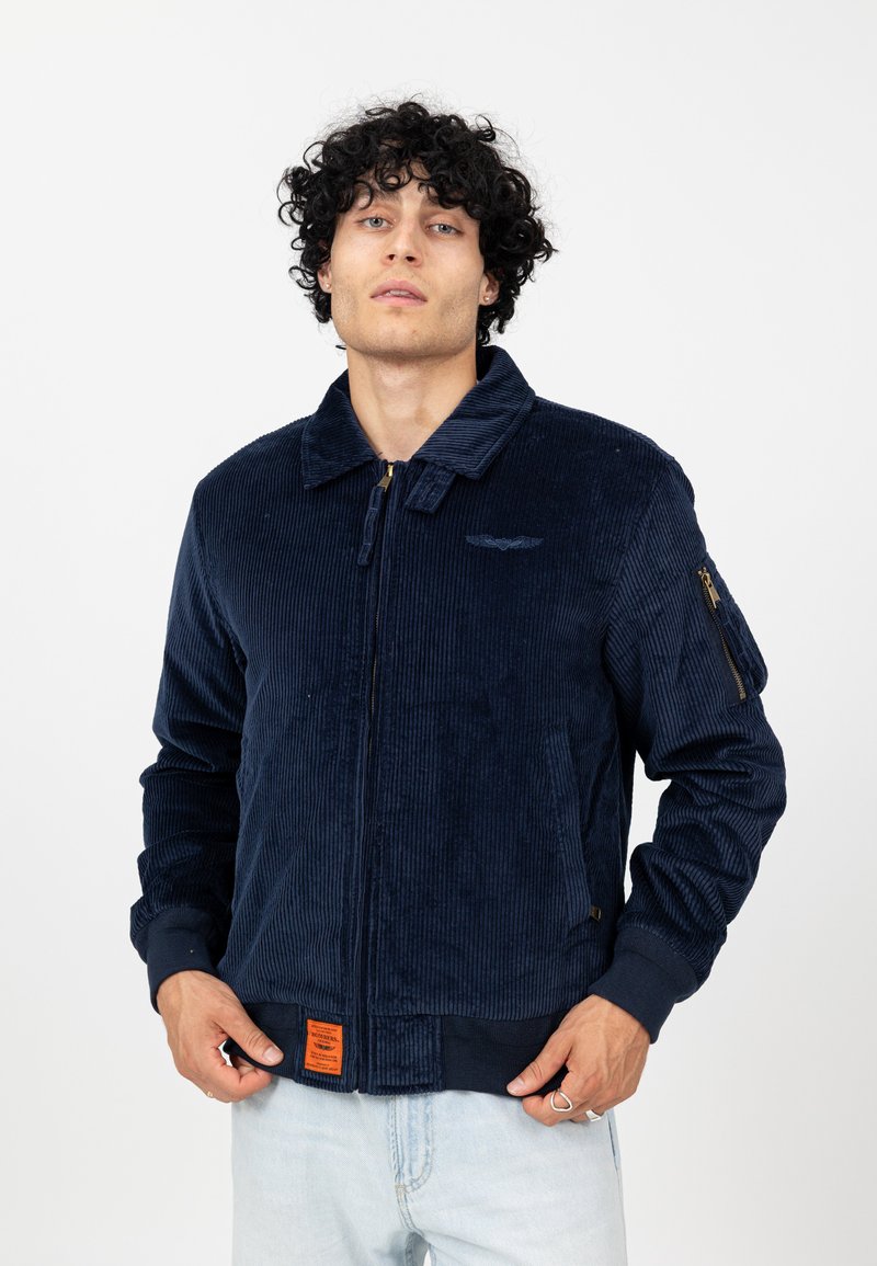 Marineblauwe corduroy jas met een ritssluiting aan de voorkant, opstaande kraag, twee zakken, een oranje label aan de zoom en geribbelde manchetten. Heeft een subtiele logo op de borst.