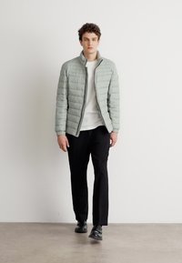 Light green quilted jacket with a high collar, worn over a white shirt and black pants. Model stands against a plain background.