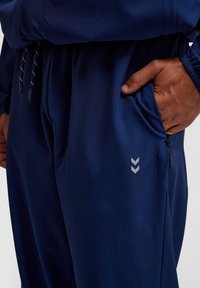 Marina atletiska byxor gjorda av slät tyg, med en dold ficka med dragkedja och en reflekterande chevron-logotyp på sidan.