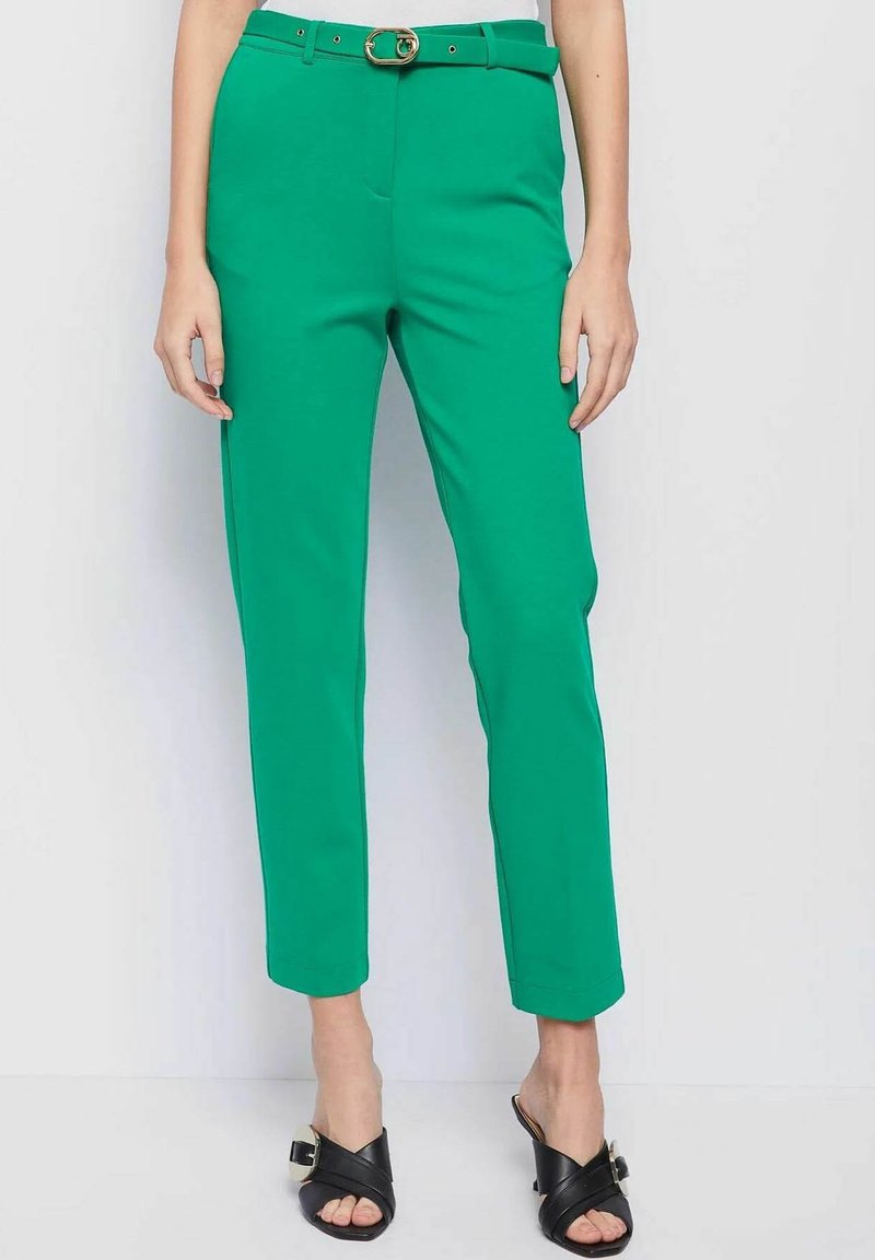 Groene, op maat gemaakte broek met een riem met een gouden gesp, taps toelopende benen, gladde textuur en zijzakken, gecombineerd met zwarte sandalen.