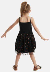 Schwarzes Kleid mit taillierter Körperschneidung und knielangem Rock. Der Rock hat kupferfarbene Sternmuster und eine durchsichtige Überlage. Braune Schuhe.