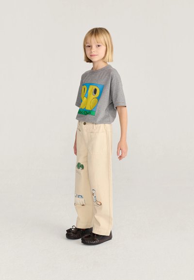 Enfant aux cheveux blonds courts, debout, portant un t-shirt graphique gris, un pantalon crème avec des patchs colorés et des chaussures marron foncé.