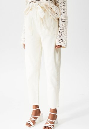 Woman wearing high-waist cream pants with a fabric belt, paired with a crochet beige sweater and white strappy heels.