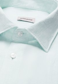 Chemise en lin bleu clair avec col pointu et boutons blancs, marquée d'une étiquette "seidensticker" cousue à l'intérieur du col.
